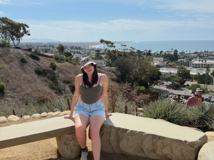 A woman in shorts and a hat poses for a photo near the ocean.