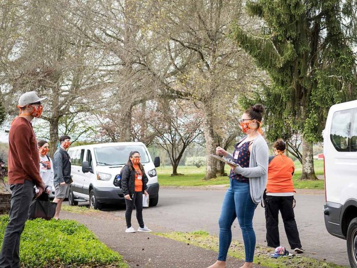 TRACE staff wearing face masks and face shields brief community volunteers before COVID-19 testing outside near two white vans.