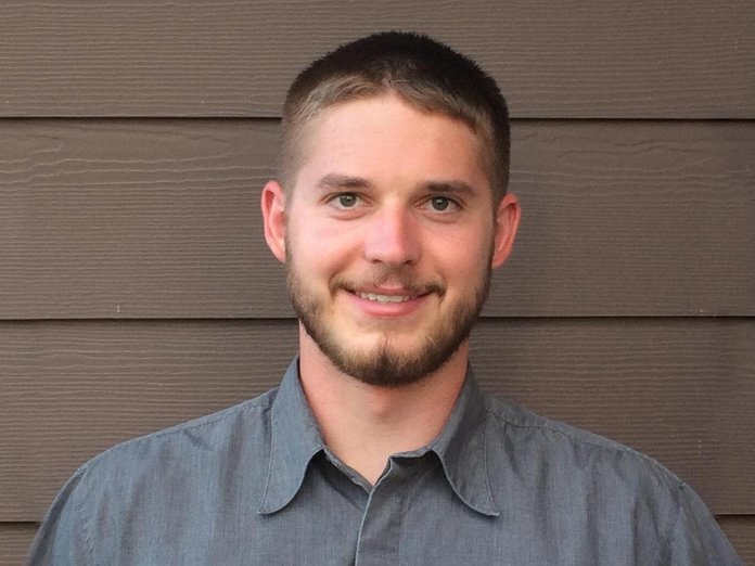A young man in a button up shirt stands in front of a house
