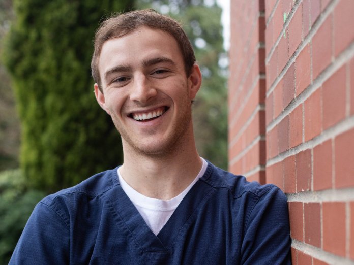 A smiling young man in scrubs leans against a brick wall