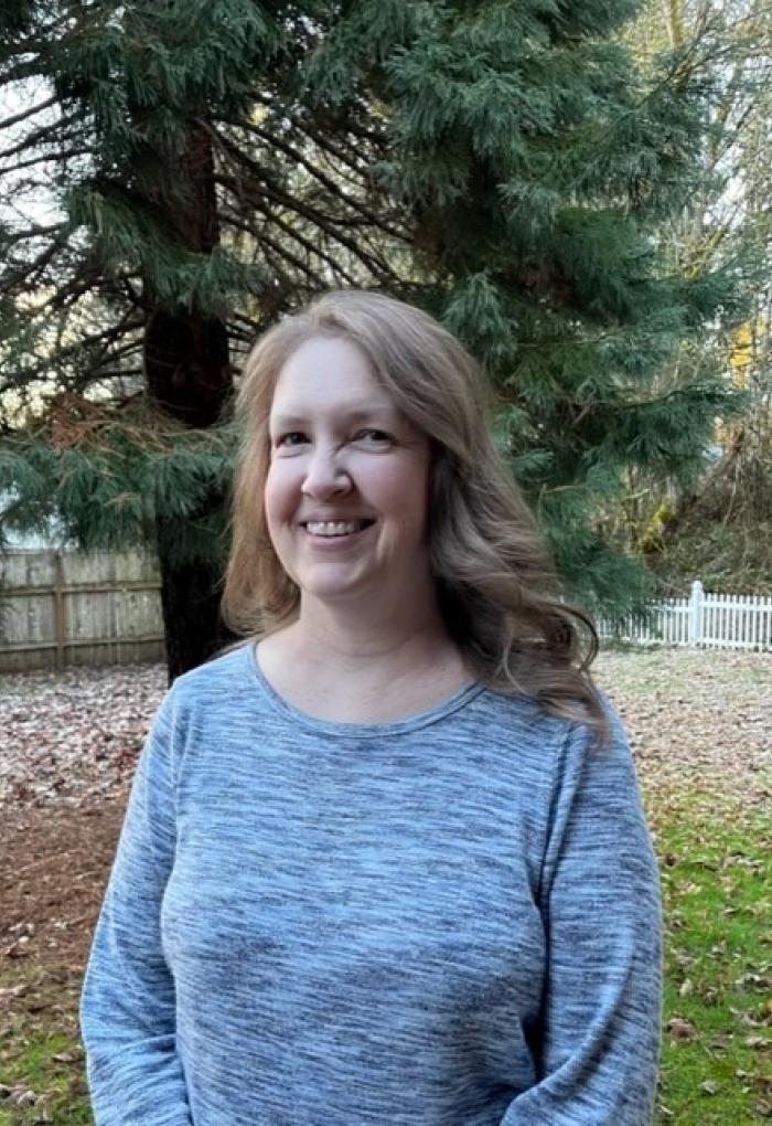 Kelly Carter standing in a blue long sleeve shirt outside in front of a tree. 