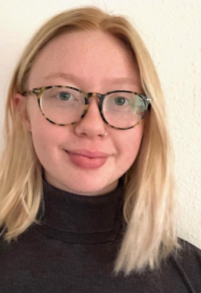 Sunni Patton standing in front of a white wall wearing a black turtle-neck and glasses.