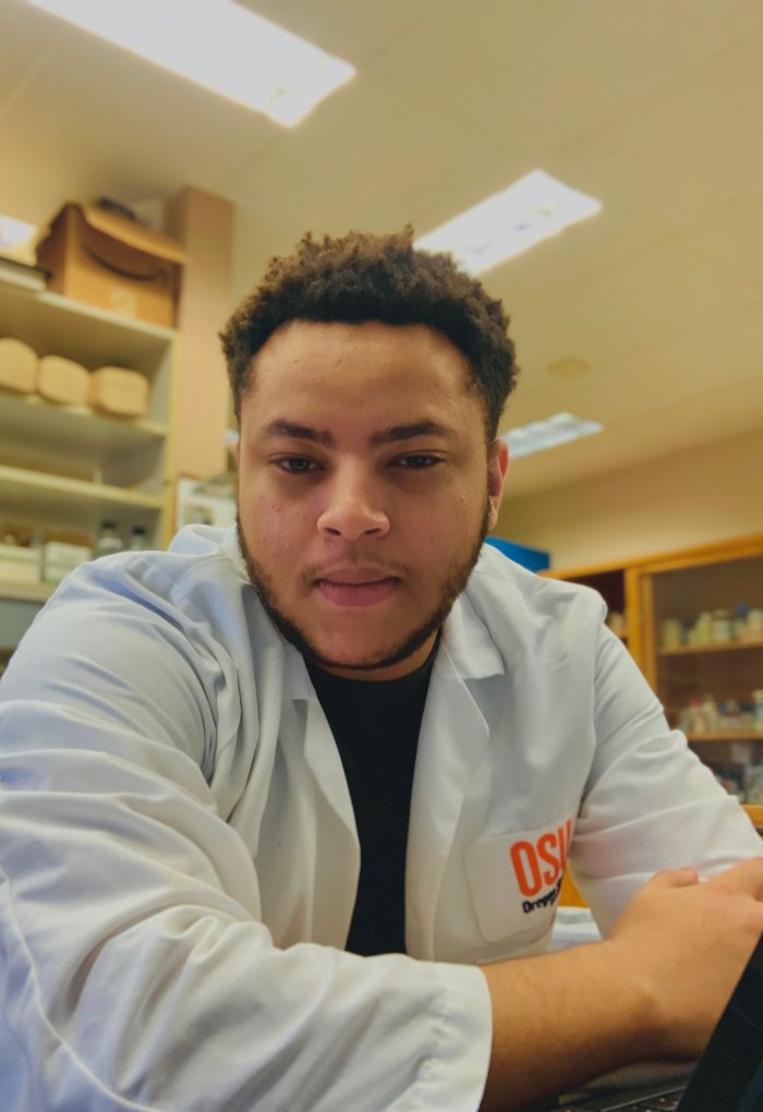 A man in an Oregon State University lab coat poses for a picture inside of a laboratory.
