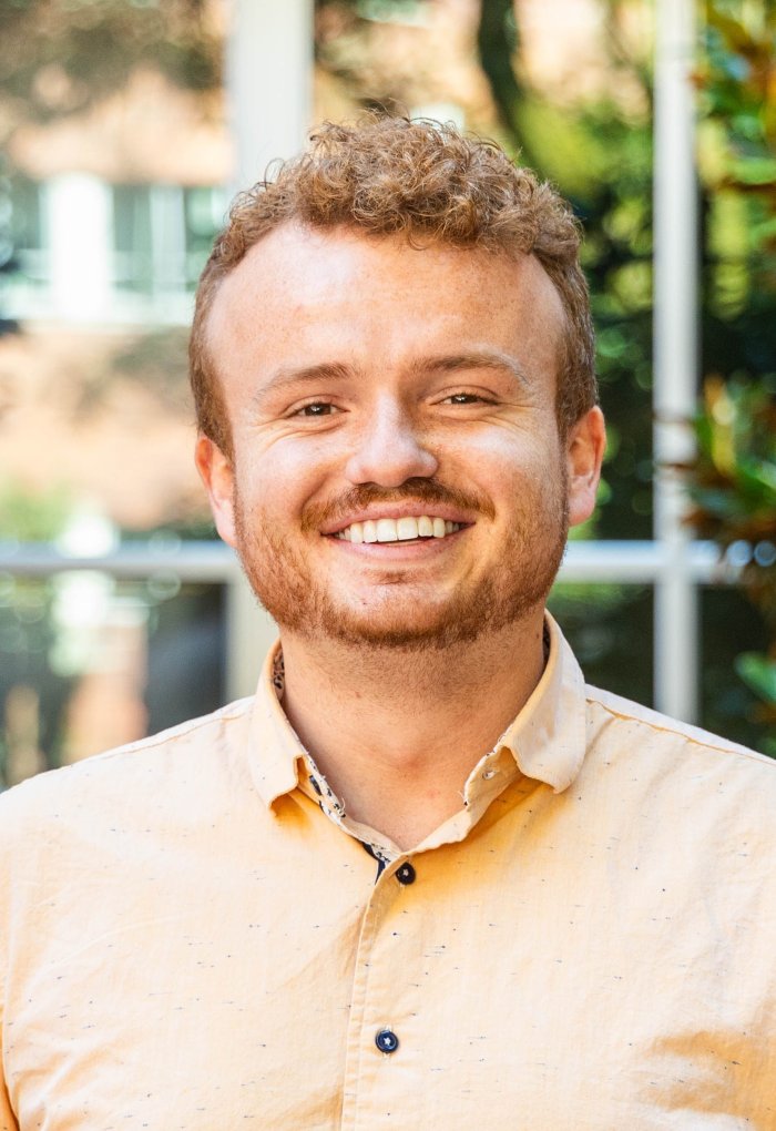 Head shot of a man with a yellow shirt in front of green trees.