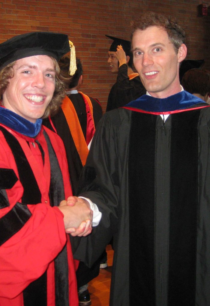 A man in a cap and gown shakes the hand of his Ph.D. advisor.