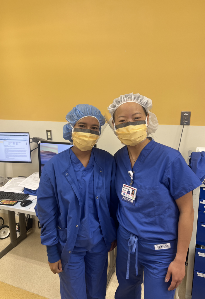 Maria Ruiz Cortes and Dr. Weiya Wysham stand side-by-side in a medical center; wearing blue scrubs and yellow masks that cover their smiles for the camera.
