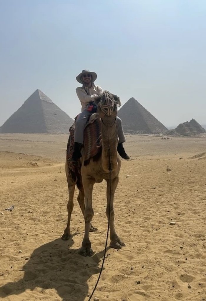 A man poses for a photo on a camel with a pyramid in the background.