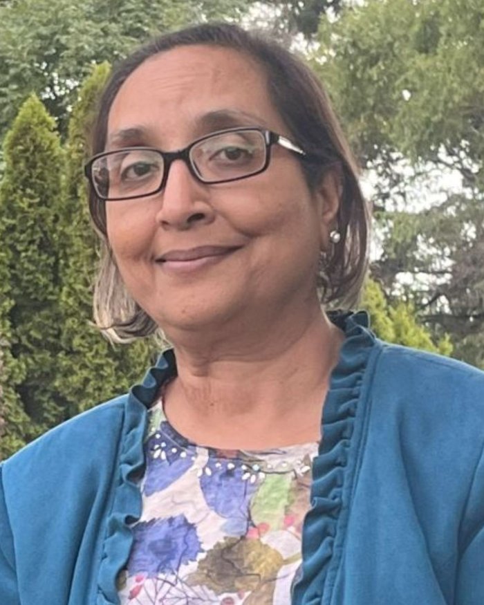 Person wearing glasses and a blue jacket over a floral-patterned top, standing outdoors in front of dense green trees and shrubs in a garden-like setting.