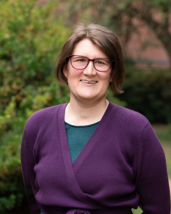 Lauren Dalton smiles outside in a purple wrap sweater. She wears glasses and has short, brown hair.