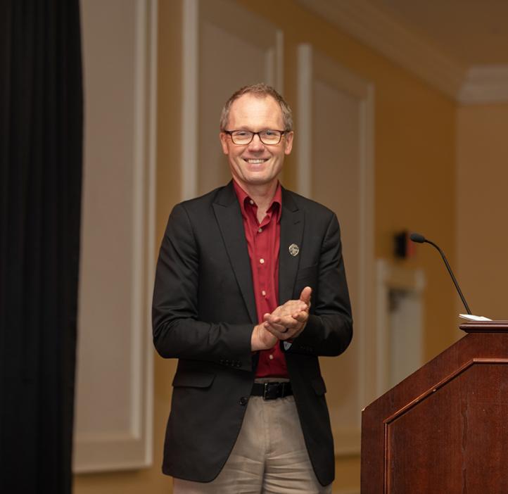 Roy standing at a podium smiling