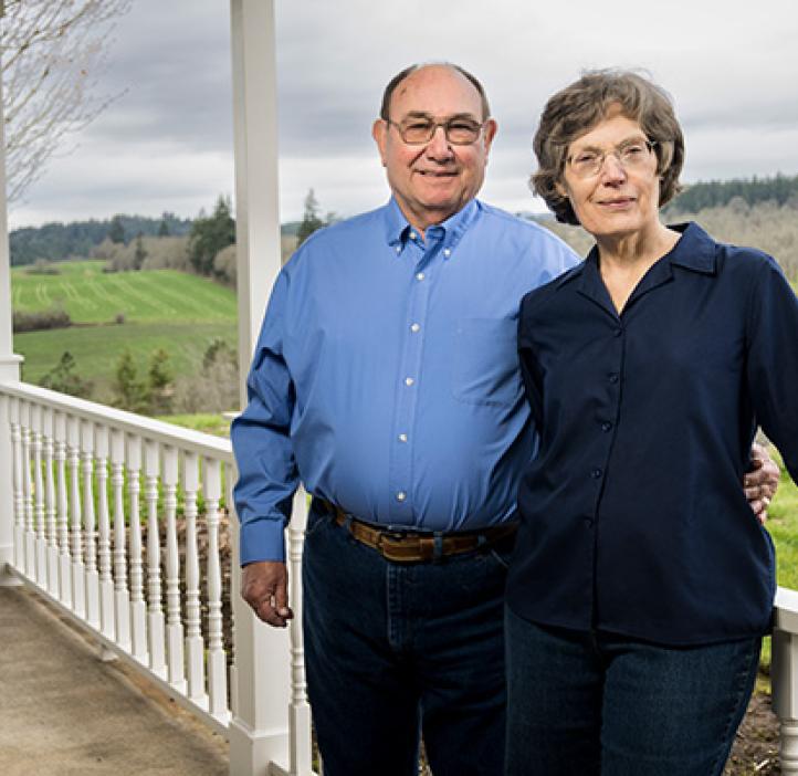 Norbert and Eileen Hartmann lean against a porch railing with green landscape behind them.