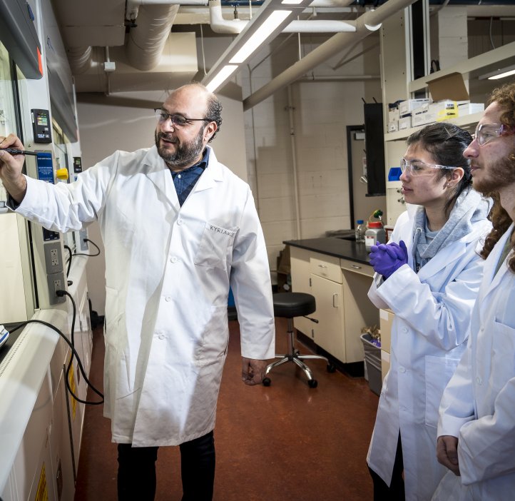 Three people in lab coats write on a clear board inside of a laboratory.