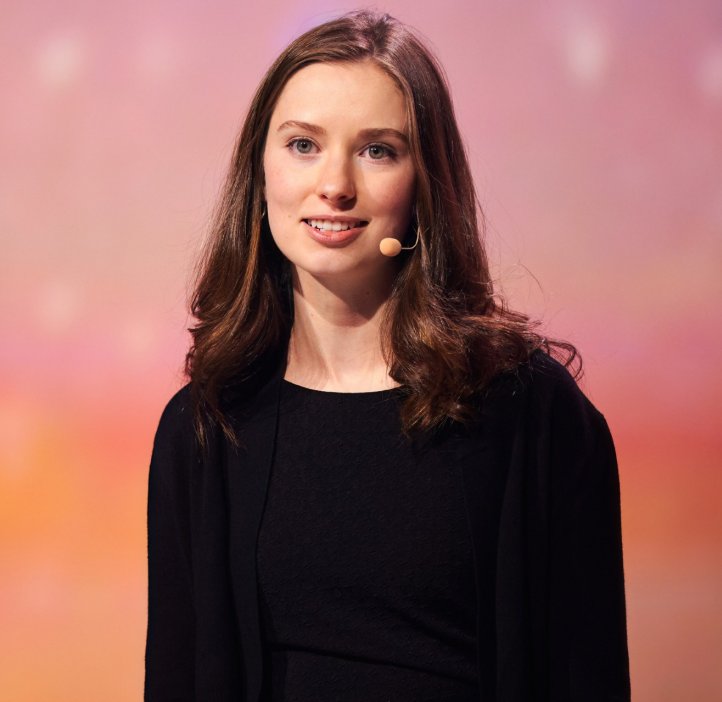 A woman with shoulder-length hair wears an earset microphone and presents to an unseen audience