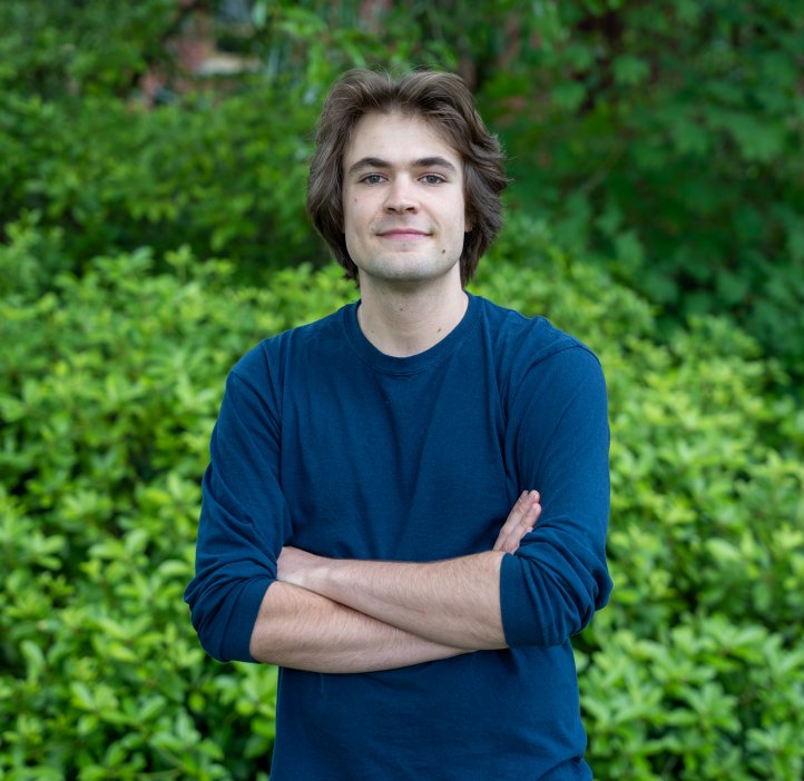 Joey Takach posing with a smile in front of leafy green bushes.