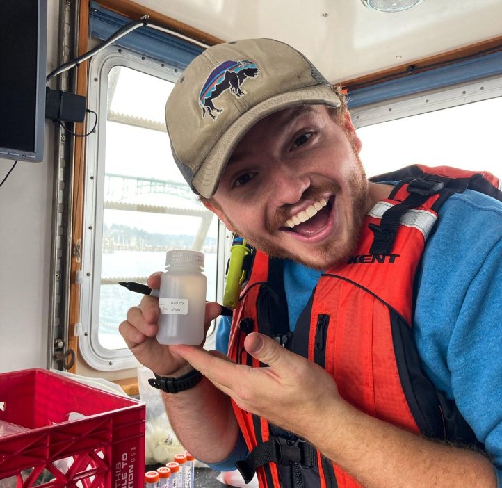 A man with a slight beard wearing a tan hat with a bison on it, and wearing an orange lifevest, holds a sample bottle.