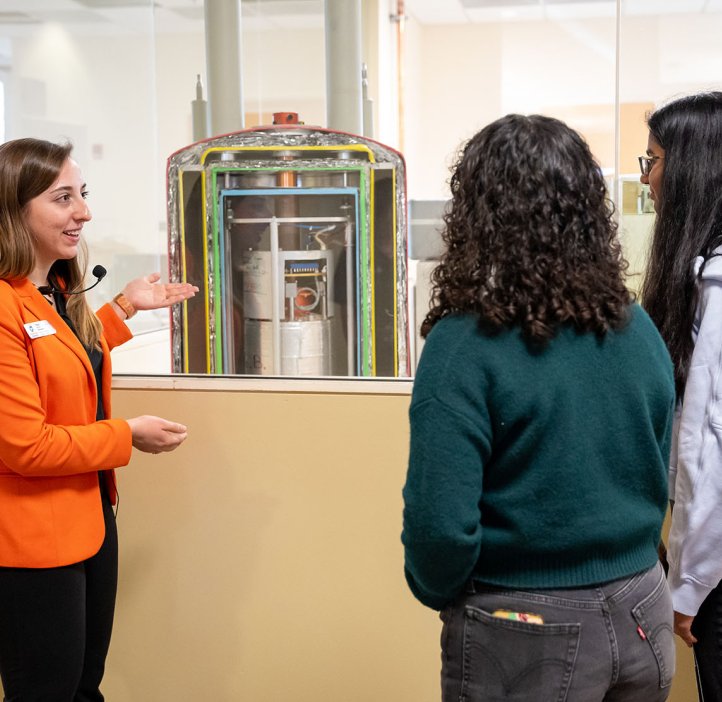 An OSU science ambassador in a vibrant orange blazer showing her two female tour guests a cross-section visual of an instrument in OSU's nuclear magnetic resonance facility.