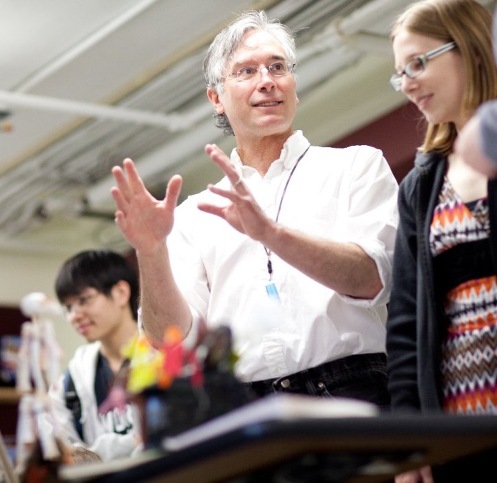 A man in a white button-up gestures to molecular art on a table, explaining it to onlooking students.