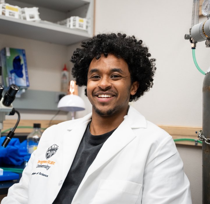 An undergraduate researcher wears an Oregon State lab coat in the lab, surrounded by equipment including a microscope and vials.