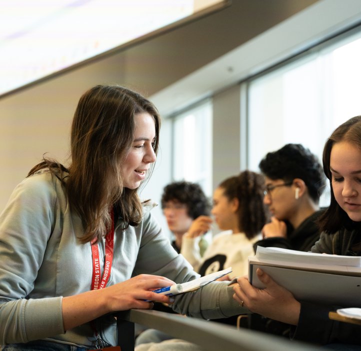 A woman leans over seat railing to aid a student, pointing to the student's notebook mid-conversation.