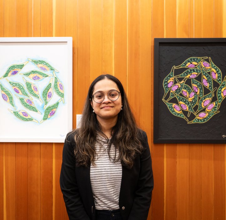 Vaishnavi Padaki stands in Strand Gallery. Two of her art pieces hang on either side of her on the wall. The one on the left is on a white background, the one on the right is on a black background. She wears glasses and has long dark hair. She smiles slightly.