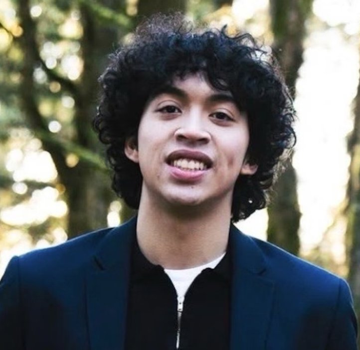 Jacob Van stands in a forest. He is visible from the chest, up wearing a black zip-up hoodie and a navy blue blazer. He has brown skin and black curly hair. He is smiling.