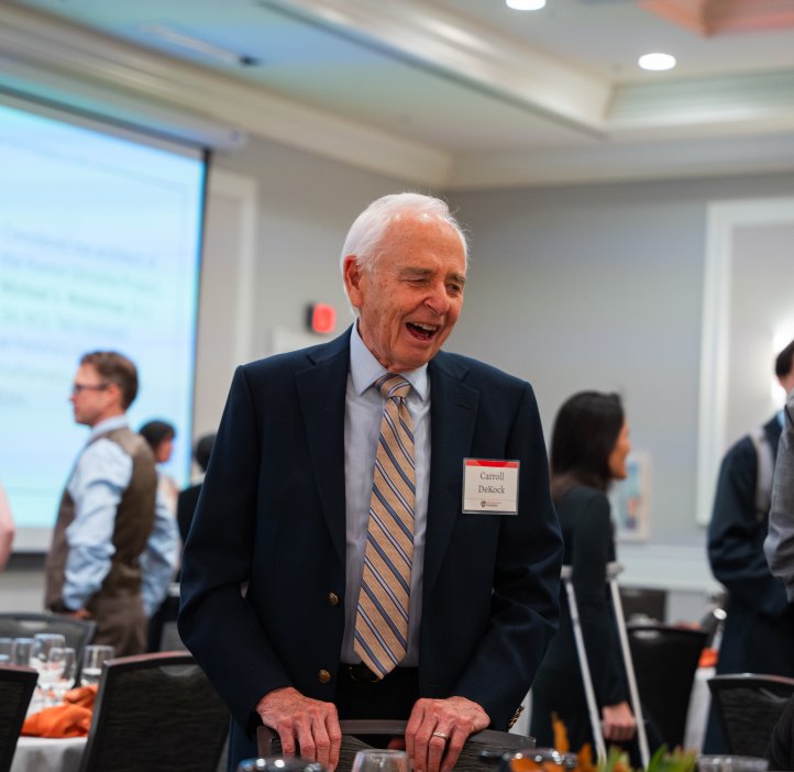 A man wearing a suit and tie smiles while talking with other guests at a table.