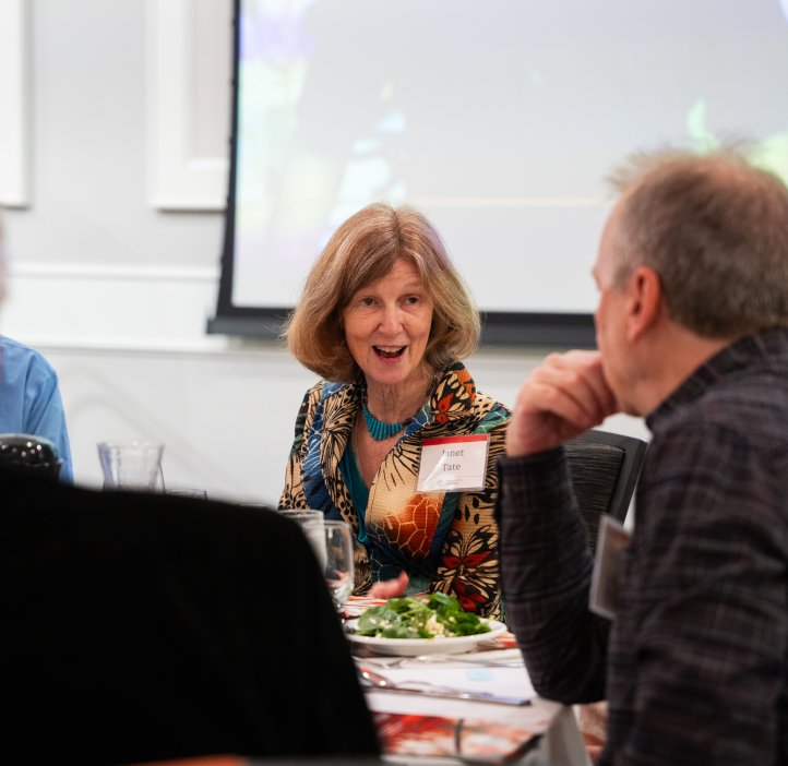 A woman with a name tag saying "Janet Tate" sits at a table and smiles while talking with others.