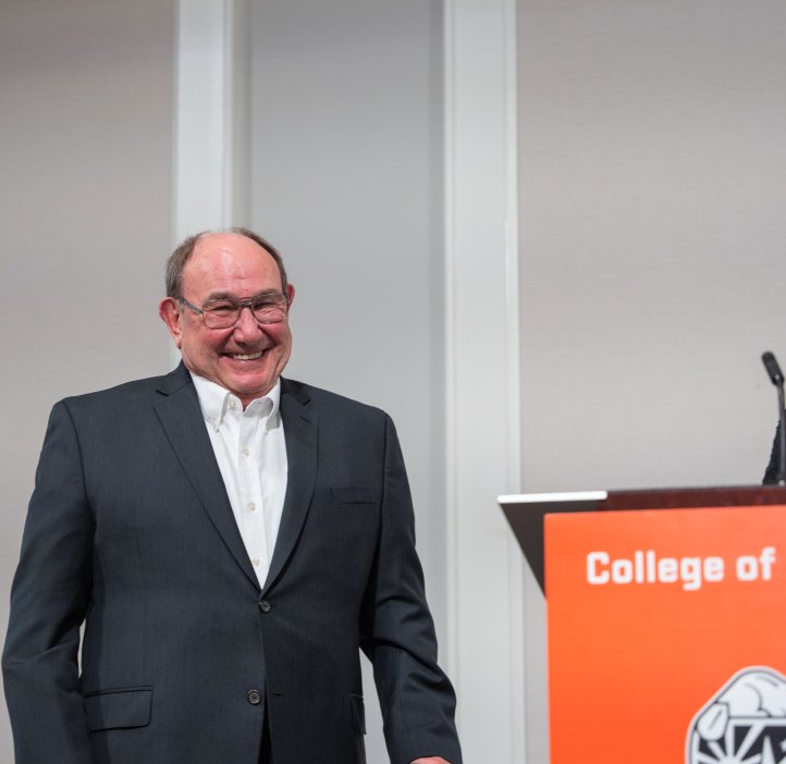 A man in a dark suit jacket and white shirts smiles at a woman wearing a red shirt speaking at a podium.