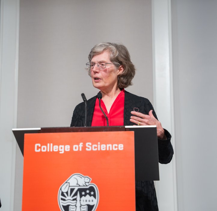 In red attire, Eileen Hartmann speaks at the College of Science Alumni Awards Ceremony from a branded podium.