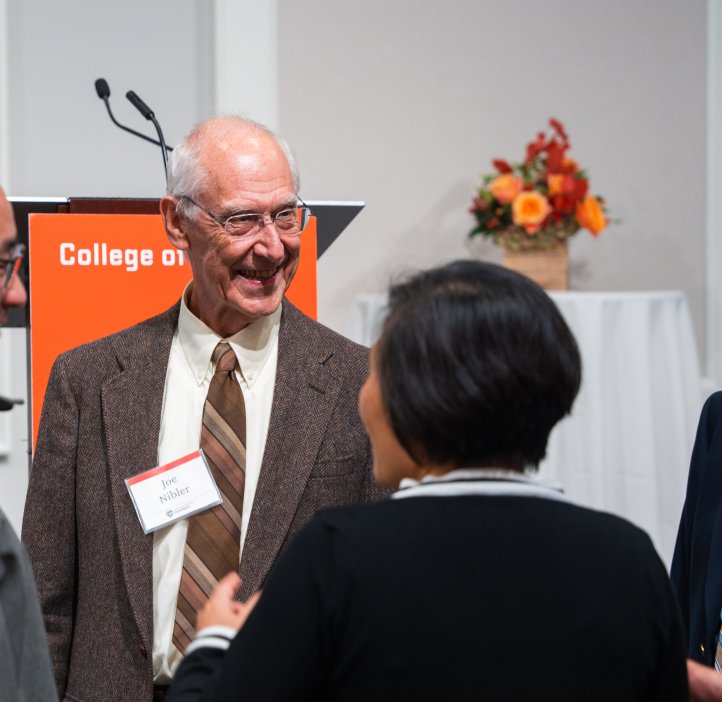 Group of attendees conversing near a podium with the College of Science logo and floral arrangement in the background.