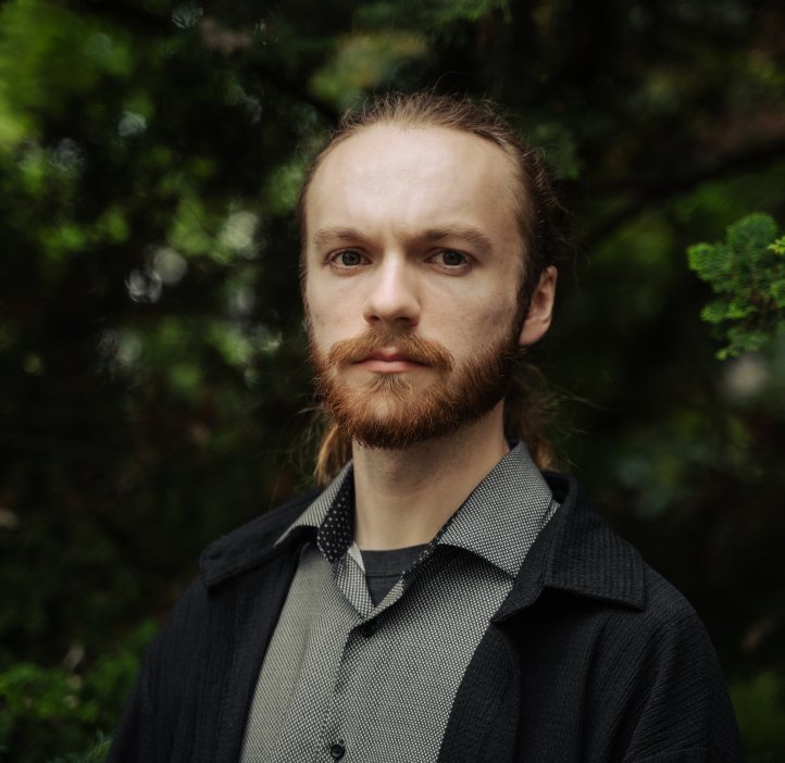 A man in a grey shirt and black coat poses for a photo in front of trees.