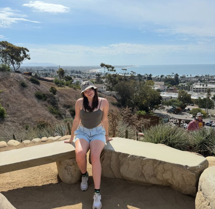 A woman in shorts and a hat poses for a photo near the ocean.