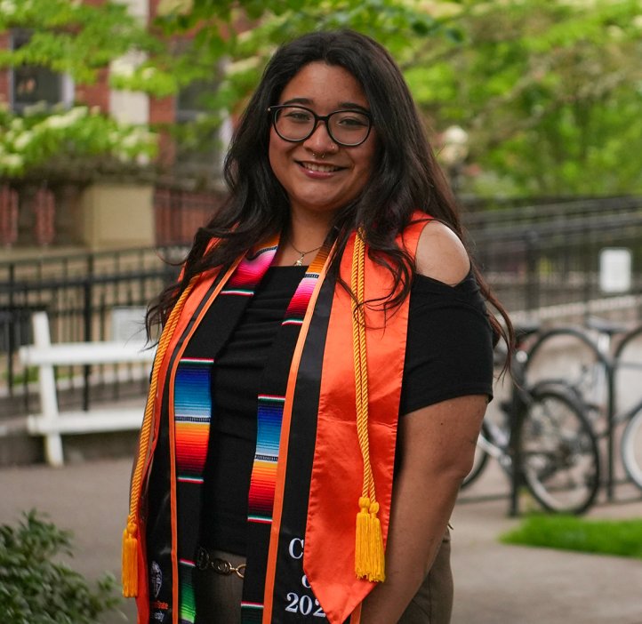 A woman in glasses and graduation regalia smiles brightly in front of a sunny campus backdrop.