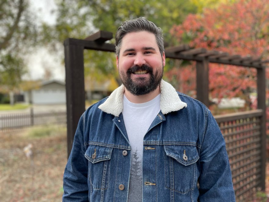 A man with a beard stands in a denim jacket with autumn colored trees in the background.