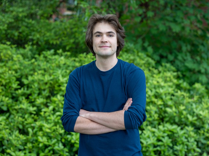 Joey Takach posing with a smile in front of leafy green bushes.