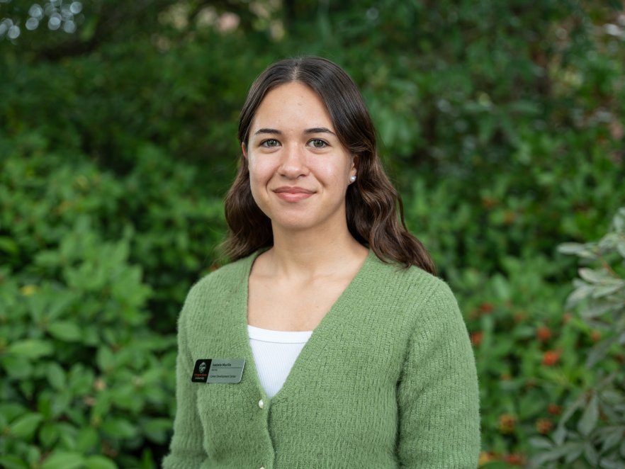 Isabella Murillo wears a green cardigan and stands in front of greenery smiling slightly. She has long brown hair.