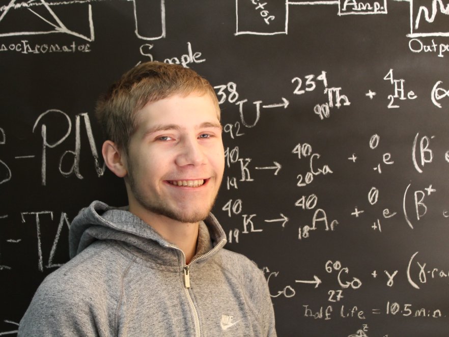 A young man in a sweatshirt stands in front of a chalkboard with many chemistry equations